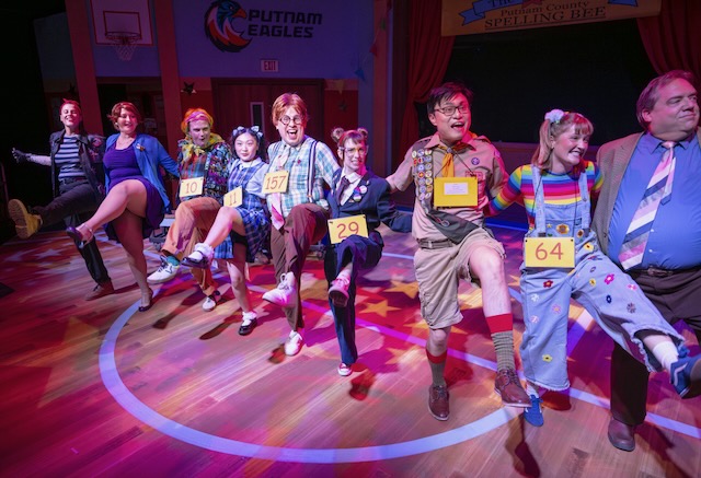 The ensemble cast performs a synchronized kick line during The 25th Annual Putnam County Spelling Bee, dancing together on a brightly lit gymnasium stage.