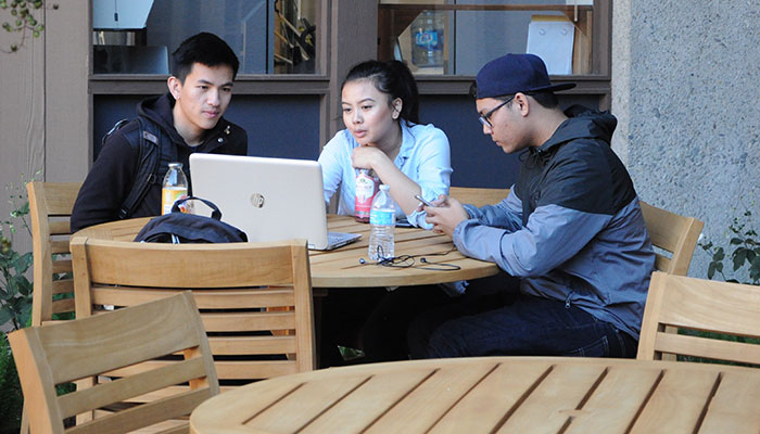 group of three students sitting at outdoor wooden tables