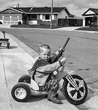 young boy holding gun on a tricycle with suburban house in background