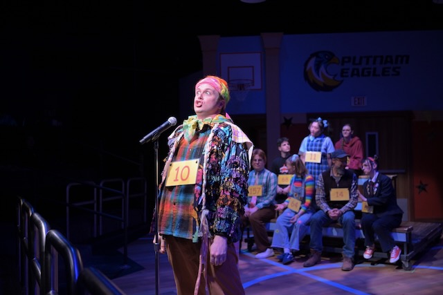 A contestant wearing number 10 stands at a microphone during a spelling bee while other competitors sit behind them on bleachers on a school gymnasium stage set.