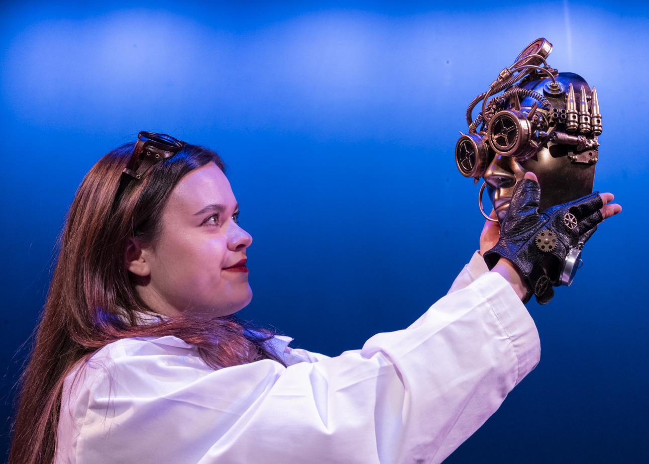 An actor in a white lab coat holds up a steampunk-style mechanical head, wearing goggles and gears, against a blue-lit background.
